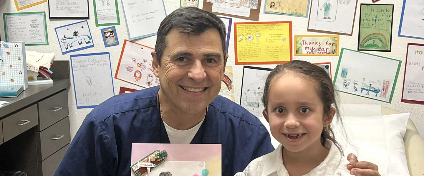 Dr. Arturo Bonilla, pediatric microtia surgeon, smiling with a young microtia patient in his San Antonio office surrounded by patient thank-you letters and drawings