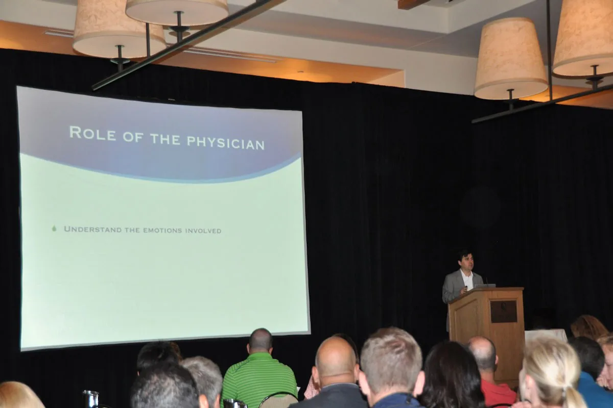 Dr. Arturo Bonilla speaks from a podium to a packed audience at a microtia conference, with a large projection screen displaying the slide Role of the Physician — Understand the Emotions Involved. The presentation reflects Dr. Bonilla's commitment to the emotional as well as surgical aspects of caring for microtia patients and their families.