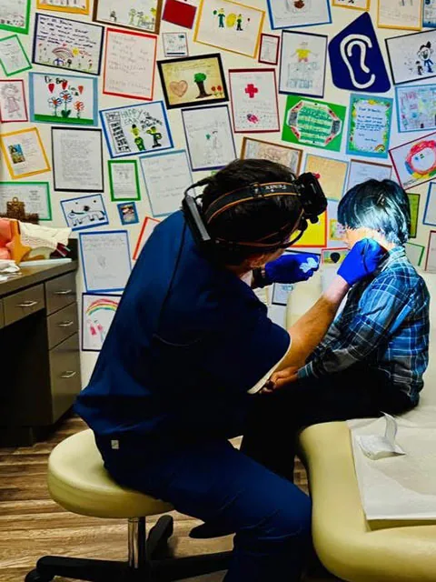 Dr. Arturo Bonilla wearing a surgical headlamp and blue gloves performs a close-up ear examination on a seated patient in his San Antonio office.