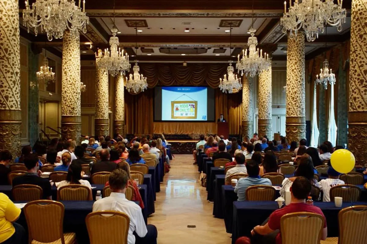 A grand ballroom with crystal chandeliers is filled with rows of families attending a microtia conference, with a presenter at the podium and a Kids Only slide displayed on the large projection screen. Dr. Arturo Bonilla organizes and speaks at microtia family conferences bringing together patients, parents, and specialists from across the country.