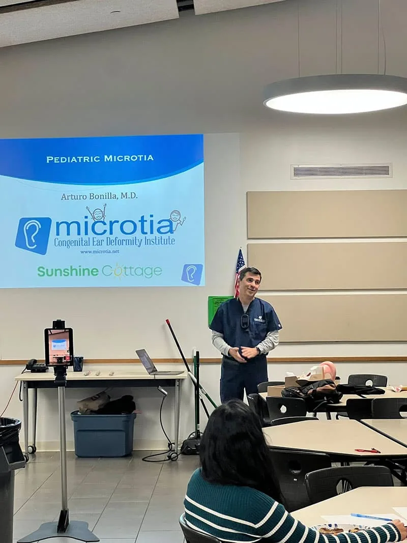 Dr. Arturo Bonilla stands in front of a projected slide reading Pediatric Microtia alongside the Microtia Congenital Ear Deformity Institute and Sunshine Cottage logos while presenting to an audience in a classroom setting. Dr. Bonilla partners with Sunshine Cottage School for the Deaf in San Antonio to educate staff about pediatric microtia and hearing loss.