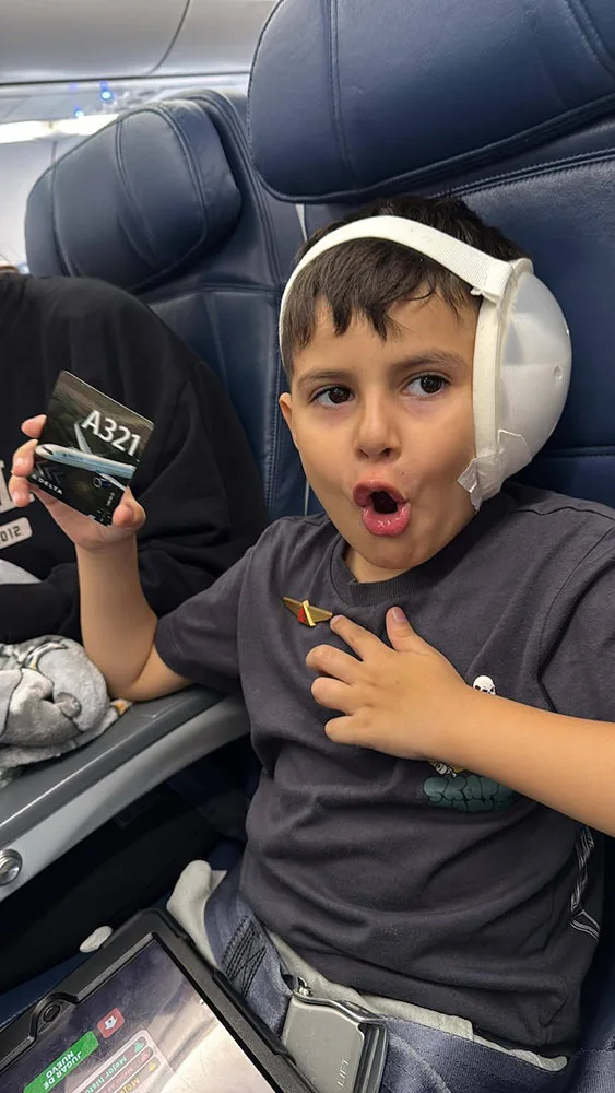 A young boy wearing a post-surgical ear protective cup sits excitedly in a Delta Airlines A321 cabin seat, holding a plane card and making a surprised expression. He is traveling to San Antonio to receive microtia surgery with Dr. Arturo Bonilla.