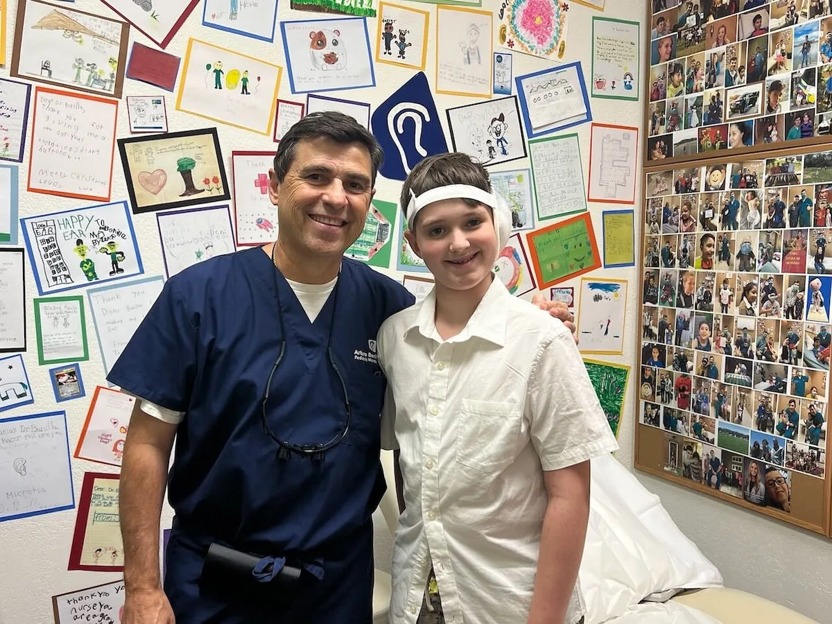 Dr. Arturo Bonilla in blue surgical scrubs stands smiling with a young boy wearing a post-surgical ear bandage in front of a wall covered in children's thank-you drawings and patient photos. The wall displays handwritten notes and artwork from microtia surgery patients treated at Dr. Bonilla's San Antonio practice.