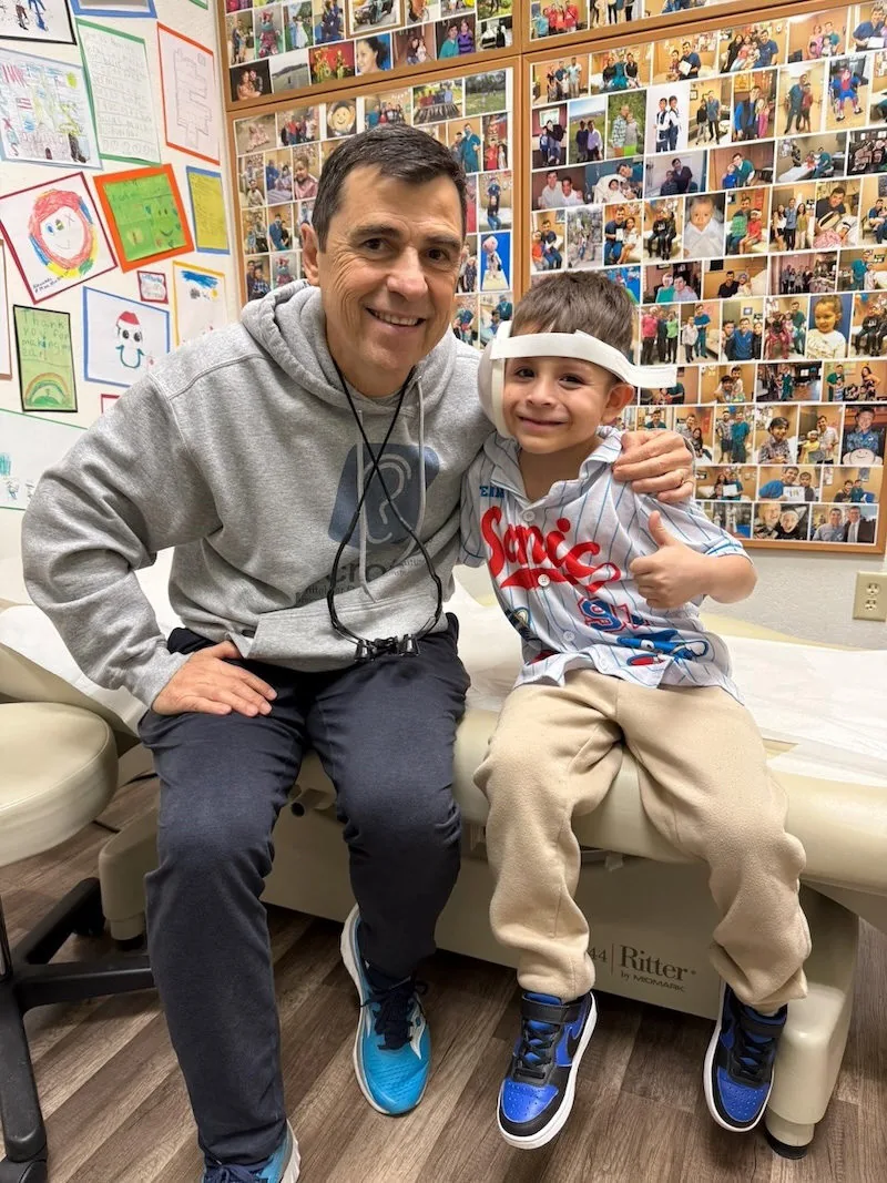 Dr. Arturo Bonilla sits beside a smiling young boy wearing a post-surgical ear bandage and giving a thumbs up in an exam room decorated with children's drawings and patient photos. The boy is a microtia surgery patient at Dr. Bonilla's San Antonio practice.