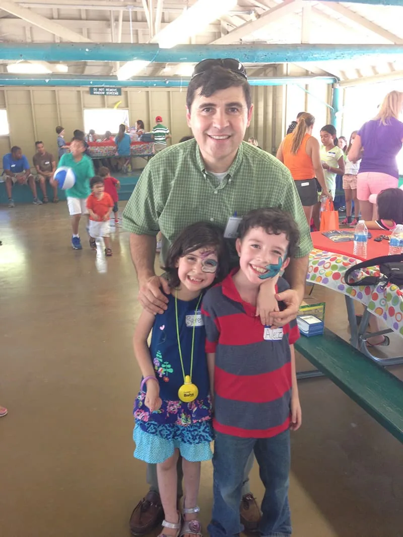 Dr. Arturo Bonilla smiles with two young face-painted children at an indoor microtia patient picnic event in San Antonio. Dr. Bonilla hosts community gatherings where microtia patients and their families can connect with one another.