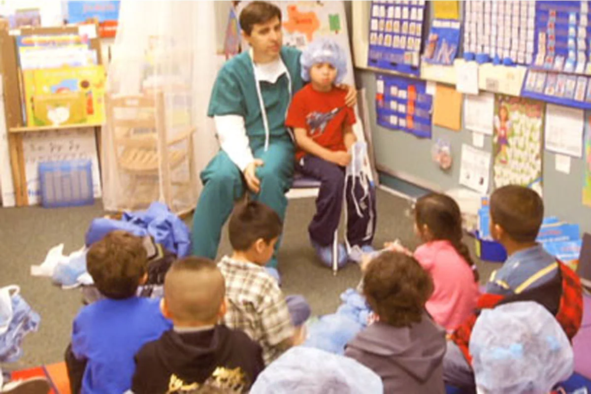 Dr. Arturo Bonilla in surgical scrubs sits beside a young microtia patient wearing a surgical cap as they speak together to a group of elementary school children seated on a classroom floor. Dr. Bonilla visits schools with his microtia patients to help classmates understand ear differences and build awareness around microtia.