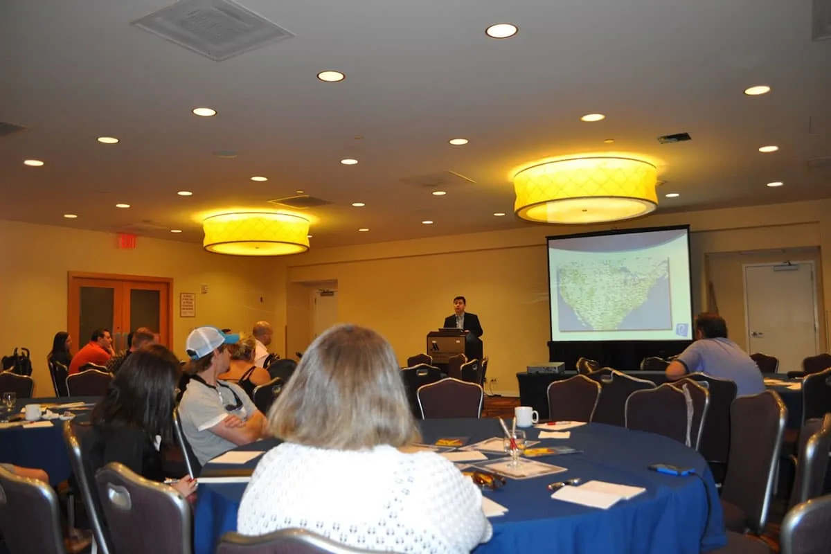 Dr. Arturo Bonilla stands at a podium presenting to a seated audience in a conference room with a large US map projected on the screen behind him. Dr. Bonilla travels to cities including El Paso to speak about pediatric microtia surgery and connect with families seeking ear reconstruction care.