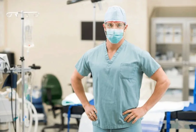 A male pediatric anesthesiologist in surgical scrubs, cap, face mask, and safety goggles stands confidently in a fully equipped operating room. Pediatric anesthesiology is a critical part of the microtia surgery team ensuring each child's safety throughout the ear reconstruction procedure.