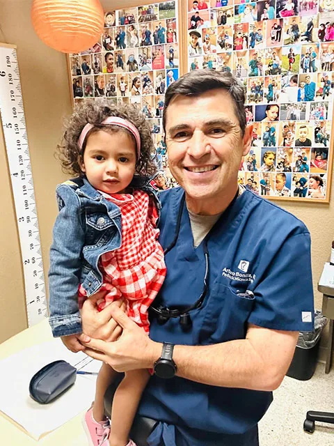 Dr. Arturo Bonilla in blue surgical scrubs smiles while holding a young toddler girl in a red plaid dress in his exam room, with a wall of patient photos visible behind them.