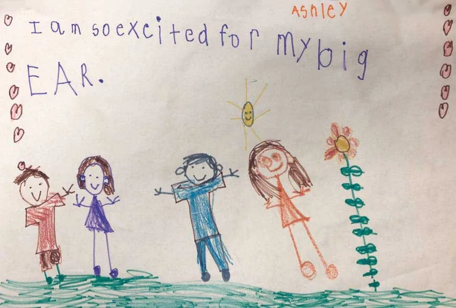 Dibujo infantil con crayones de Ashley, que muestra cuatro figuras alegres de palitos bajo un sol sonriente, con el mensaje «Estoy muy emocionada por mi gran OREJA»: un entusiasta regalo de agradecimiento al Dr. Arturo Bonilla de una paciente agradecida con microtia que espera su reconstrucción de oreja