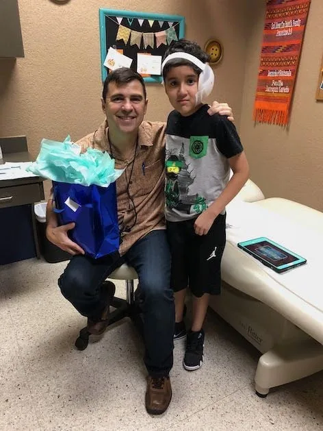 Fotografía del Dr. Arturo Bonilla arrodillado junto a un paciente joven con microtia que lleva un vendaje postoperatorio en la cabeza, mientras el doctor sostiene una bolsa de regalo azul entregada durante la visita al consultorio: un generoso momento de agradecimiento al Dr. Arturo Bonilla de un paciente agradecido que se sometió a cirugía de microtia