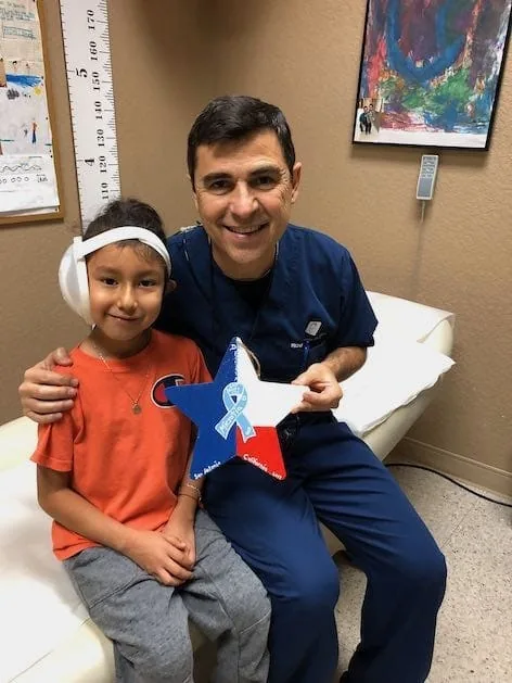 Fotografía del Dr. Arturo Bonilla con ropa quirúrgica azul, sonriendo con un paciente joven vendado con microtia que sostiene un adorno de estrella pintada con un lazo de concientización sobre la microtia e inscripciones de San Antonio y California: un orgulloso momento de entrega de un regalo después de la cirugía con un paciente agradecido que se sometió a reconstrucción por microtia