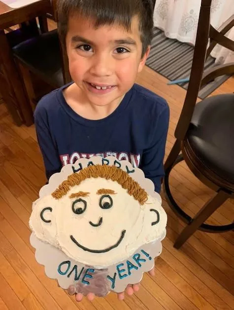 Fotografía de un niño sonriente que sostiene un pastel decorado con un diseño de rostro y el texto «Un año», para celebrar un año desde su reconstrucción de oreja por microtia: un alegre regalo que conmemora este momento al Dr. Arturo Bonilla de un paciente agradecido que se sometió a cirugía de microtia
