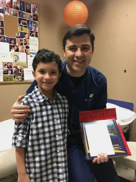 Fotografía del Dr. Arturo Bonilla con ropa quirúrgica azul marino, sonriendo con un paciente joven con microtia que le regaló un álbum de fotografías colorido: un cálido momento de agradecimiento en el consultorio capturado como regalo al Dr. Arturo Bonilla de un paciente agradecido que se sometió a cirugía de microtia