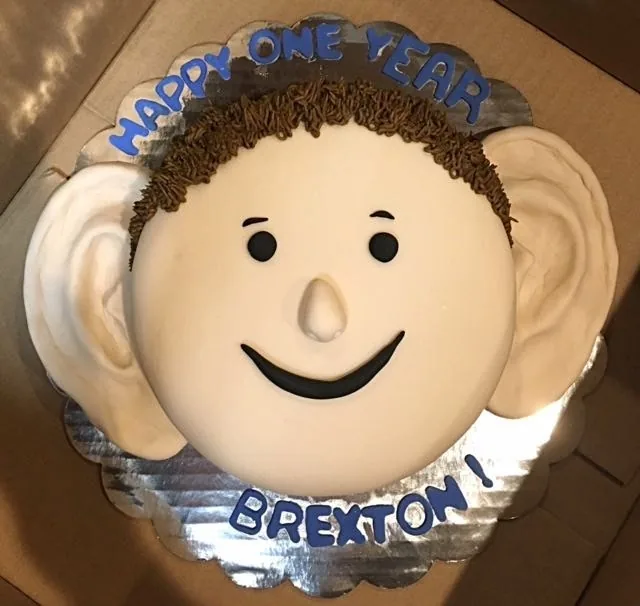 Pastel de celebración personalizado para un paciente llamado Brexton, con el texto «Feliz primer año, Brexton», decorado como una cara sonriente con dos orejas esculpidas: un alegre regalo que conmemora el primer año de la reconstrucción por microtia y honra la cirugía de oreja del Dr. Arturo Bonilla que cambia vidas
