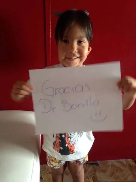 Fotografía de un niño pequeño que sonríe y sostiene con orgullo un cartel escrito a mano que dice «Gracias Dr. Bonilla», con una cara sonriente: un alegre agradecimiento en español al Dr. Arturo Bonilla de un paciente agradecido que se sometió a cirugía de microtia