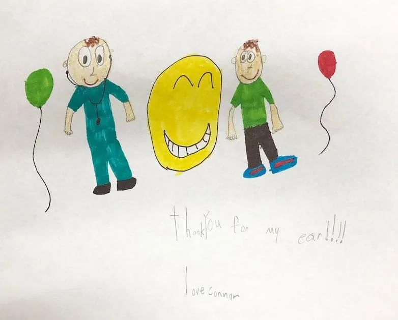 Dibujo infantil con marcadores de Connor, que muestra una figura con ropa quirúrgica verde azulada y un niño sosteniendo globos a ambos lados de una gran cara sonriente amarilla, con el mensaje «Gracias por mi oreja»: un alegre regalo de agradecimiento al Dr. Arturo Bonilla de un paciente agradecido que se sometió a cirugía de microtia