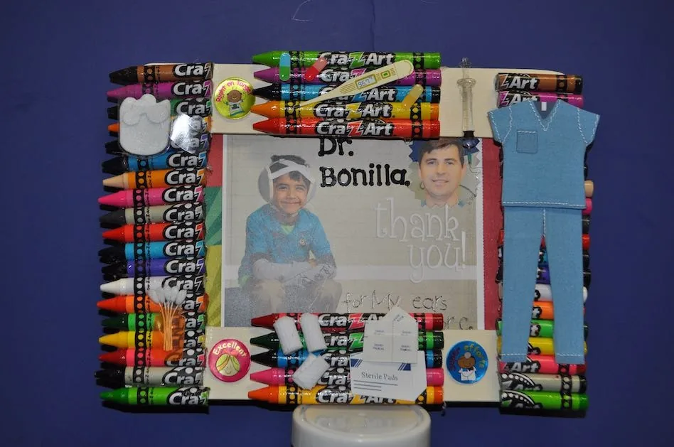 Marco hecho a mano decorado con filas de crayones y calcomanías médicas que rodean una fotografía de un paciente con microtia junto al Dr. Arturo Bonilla y un mensaje de «gracias por mis orejas»: un regalo creativo y emotivo al Dr. Arturo Bonilla de un paciente agradecido que se sometió a cirugía de microtia