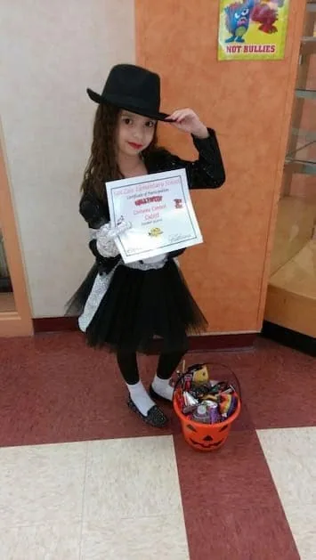 Fotografía de una niña con un disfraz negro de Halloween, sosteniendo un certificado de participación en un concurso de disfraces y un recipiente de calabaza para dulces: una alegre fotografía de un momento importante de su vida, compartida con el Dr. Arturo Bonilla por una paciente agradecida que se sometió a cirugía de microtia y celebra experiencias normales de la infancia después de la reconstrucción de oreja