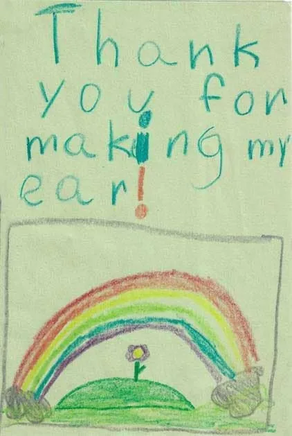 Dibujo infantil con crayones sobre papel verde, con un arcoíris colorido y una flor, y un gran texto escrito a mano que dice «Gracias por hacerme la oreja»: un alegre regalo al Dr. Arturo Bonilla de un paciente joven y agradecido que se sometió a cirugía de microtia