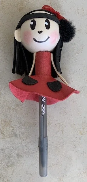 Adorno artesanal para lápiz con forma de muñeca de gomaespuma, de cabello negro y vestido rojo, regalado al Dr. Arturo Bonilla como muestra de aprecio de la familia de un paciente agradecido que se sometió a cirugía de microtia