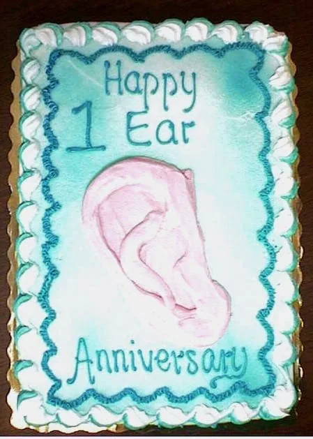 Pastel rectangular decorado con el texto «Feliz 1.er aniversario de la oreja» en glaseado verde azulado y una oreja rosa esculpida en el centro: un pastel de celebración regalado al Dr. Arturo Bonilla para conmemorar un año desde la cirugía de reconstrucción de oreja por microtia de un paciente