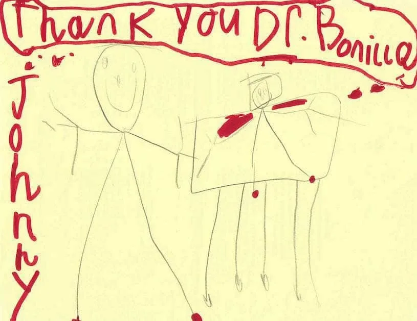 Tarjeta amarilla de un niño pequeño llamado Johnny, con «Gracias Dr. Bonilla» escrito a mano en rojo y dos figuras de palitos dibujadas a lápiz: un regalo sencillo y emotivo al Dr. Arturo Bonilla de un paciente agradecido que se sometió a cirugía de microtia