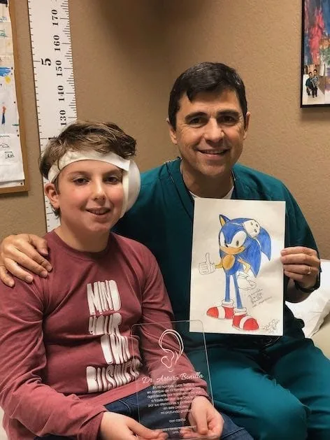 Fotografía del Dr. Arturo Bonilla con ropa quirúrgica verde azulada, sentado junto a un paciente vendado con microtia llamado Luca, que sostiene el dibujo de Sonic the Hedgehog y una placa de reconocimiento acrílica personalizada: un memorable momento de agradecimiento en el consultorio de un paciente agradecido que se sometió a reconstrucción por microtia