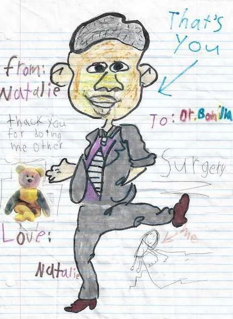 Caricatura expresiva con marcadores de una niña llamada Natalie en papel de cuaderno, dirigida al Dr. Arturo Bonilla, con una flecha rotulada «Ese es usted», junto a un agradecimiento por haberle realizado otra cirugía, un pequeño autorretrato y una calcomanía de un osito de peluche: un homenaje animado y personal de una paciente que regresa para otra cirugía de microtia