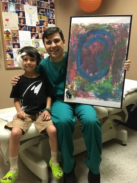 Fotografía del Dr. Arturo Bonilla con ropa quirúrgica, sentado junto a un paciente joven con microtia, ambos sonriendo mientras el Dr. Bonilla sostiene la pintura abstracta enmarcada de una oreja que le regalaron: un momento conmovedor entre el cirujano y un paciente agradecido que se sometió a reconstrucción por microtia