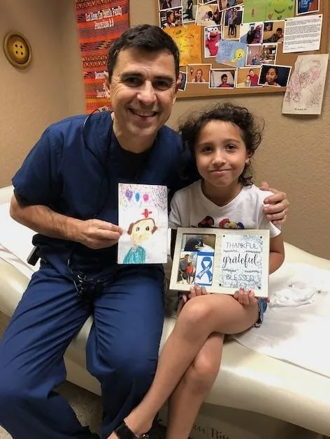 Fotografía del Dr. Arturo Bonilla con ropa quirúrgica azul marino, sentado junto a una paciente joven con microtia, ambos sosteniendo su dibujo hecho a mano y un collage fotográfico enmarcado que dice «Agradecida, llena de gratitud y bendecida», con un lazo de concientización sobre la microtia: un cálido momento de agradecimiento regalado al Dr. Arturo Bonilla por una paciente agradecida que se sometió a cirugía de microtia