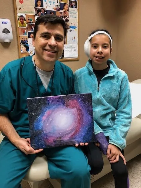 Fotografía del Dr. Arturo Bonilla con ropa quirúrgica verde azulada, sentado junto a una paciente joven con microtia que lleva una banda postoperatoria en la cabeza, ambos sonriendo mientras él sostiene una impresionante pintura acrílica de una galaxia que ella creó como regalo: un hermoso agradecimiento artístico al Dr. Arturo Bonilla de una paciente agradecida que se sometió a cirugía de microtia