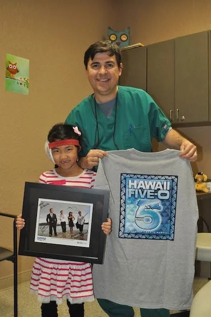 Una paciente joven de pie con el Dr. Bonilla en su consultorio, sosteniendo una fotografía enmarcada del elenco y una camiseta del programa Hawaii Five-O