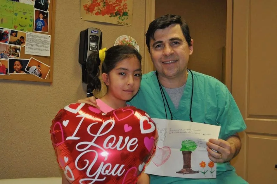 Fotografía del Dr. Arturo Bonilla con ropa quirúrgica, posando junto a una paciente joven con microtia que sostiene un globo rojo en forma de corazón con el texto «Lo quiero», mientras él sostiene su tarjeta de agradecimiento dibujada a mano: un cálido y cariñoso regalo de agradecimiento al Dr. Arturo Bonilla de una paciente agradecida que se sometió a cirugía de microtia