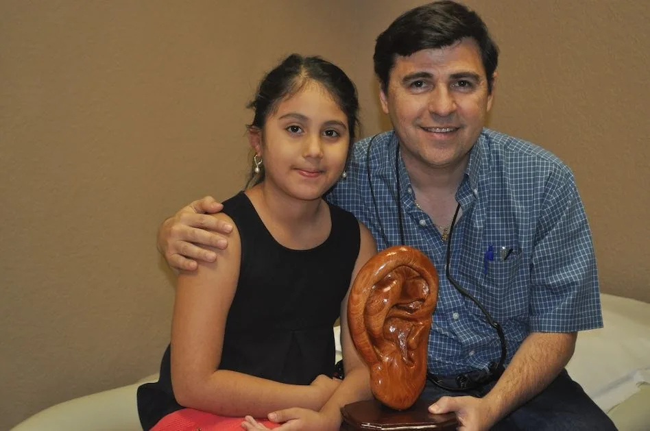 Fotografía del Dr. Arturo Bonilla sonriendo junto a una paciente joven con microtia, ambos posando con una escultura de oreja de madera pulida que le regalaron: un momento conmovedor en el consultorio que recoge la gratitud después de una cirugía exitosa de reconstrucción de oreja por microtia