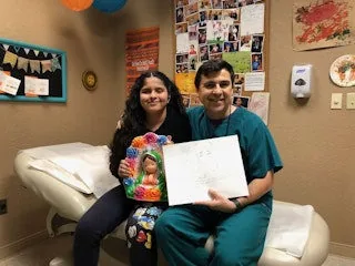 Fotografía del Dr. Arturo Bonilla sentado en su consultorio junto a un paciente con microtia que sostiene una muñeca colorida y un dibujo hecho a mano: un cálido momento de agradecimiento en el consultorio, capturado como regalo de un paciente agradecido que se sometió a reconstrucción por microtia