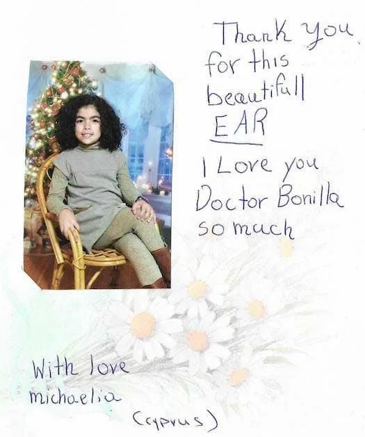 Testimonio sobre la cirugía de microtia del Dr. Bonilla, con la fotografía de una niña de Chipre junto a un árbol de Navidad y una nota escrita a mano que le agradece su hermosa oreja