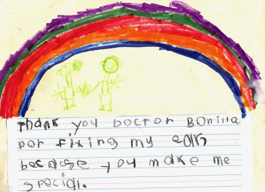 Dibujo infantil con crayones de un gran arcoíris con dos figuras de palitos y una nota escrita a mano que agradece al Dr. Arturo Bonilla haberle arreglado la oreja y haberle hecho sentir especial: un regalo emotivo de un paciente agradecido que se sometió a cirugía de microtia