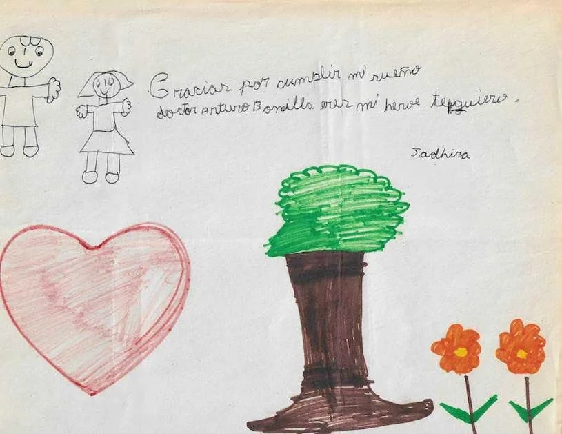 Dibujo infantil en español de Sadhira que dice «Gracias por cumplir mi sueño doctor Arturo Bonilla eres mi héroe te quiero», ilustrado con un gran corazón rojo, un árbol y flores naranjas: un agradecimiento profundamente personal al Dr. Arturo Bonilla de una paciente agradecida que se sometió a cirugía de microtia