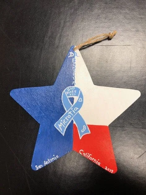 Adorno de madera en forma de estrella pintado a mano en rojo, blanco y azul, con un lazo de concientización sobre la microtia y las palabras San Antonio y California 2019: un recuerdo patriótico regalado al Dr. Arturo Bonilla por un paciente agradecido que se sometió a cirugía de microtia
