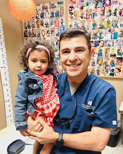 Dr. Arturo Bonilla, pediatric microtia surgeon, holds a smiling toddler with bilateral microtia in his San Antonio office, surrounded by photos of the thousands of patients he has treated from over 50 countries worldwide.