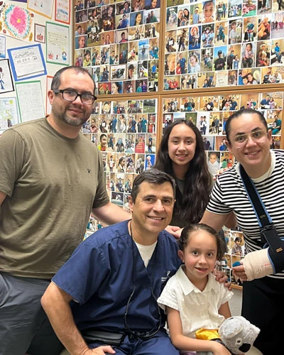 Dr. Arturo Bonilla, the world's only exclusive pediatric microtia surgeon, poses with a grateful patient family in his San Antonio office, surrounded by a wall of thousands of photos representing families he has treated from over 50 countries worldwide.
