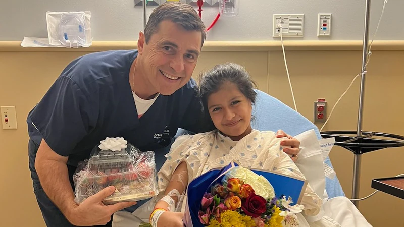Dr. Arturo Bonilla smiles with a young microtia patient in the recovery room at North Central Baptist Children's Hospital in San Antonio after successful ear reconstruction surgery — the girl holds a colorful bouquet of flowers and looks completely comfortable and happy.