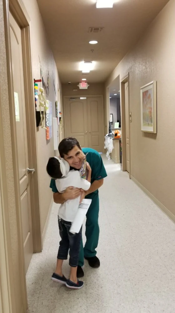 Dr. Arturo Bonilla beams with joy while a young microtia patient wraps his arms around him in a warm hug in the hallway of his San Antonio practice. The moment celebrates the successful bilateral microtia ear reconstruction Dr. Bonilla performed on both of the child's ears.
